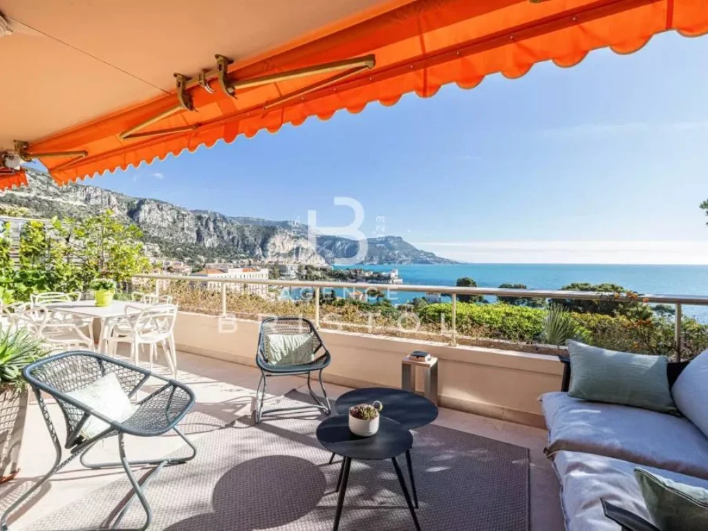 Espace extérieur cosy avec coin repas et salon sous auvent orangé, face à la mer et aux reliefs côtiers. Une ambiance chic et décontractée, idéale pour profiter de la douceur de vivre sur la Côte d’Azur.