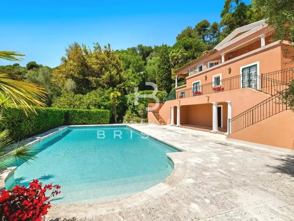 Grande villa méditerranéenne de couleur ocre, entourée de végétation luxuriante, avec une piscine turquoise et une terrasse en pierre naturelle. Un cadre paisible, élégant et ensoleillé, idéal pour une résidence haut de gamme sur la Côte d’Azur.
