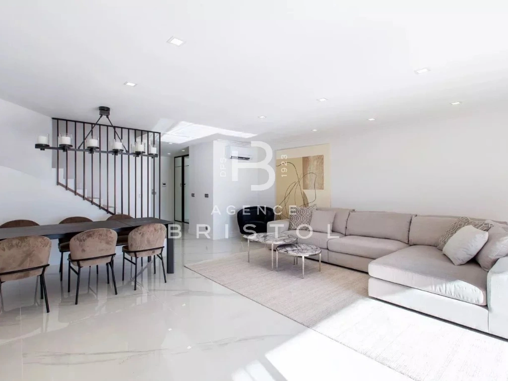 Intérieur contemporain avec sol en marbre blanc, grand canapé d’angle gris clair, table basse design et espace salle à manger avec six chaises en velours beige. Ambiance élégante, épurée et haut de gamme, idéale pour une villa de luxe sur la Côte d’Azur.