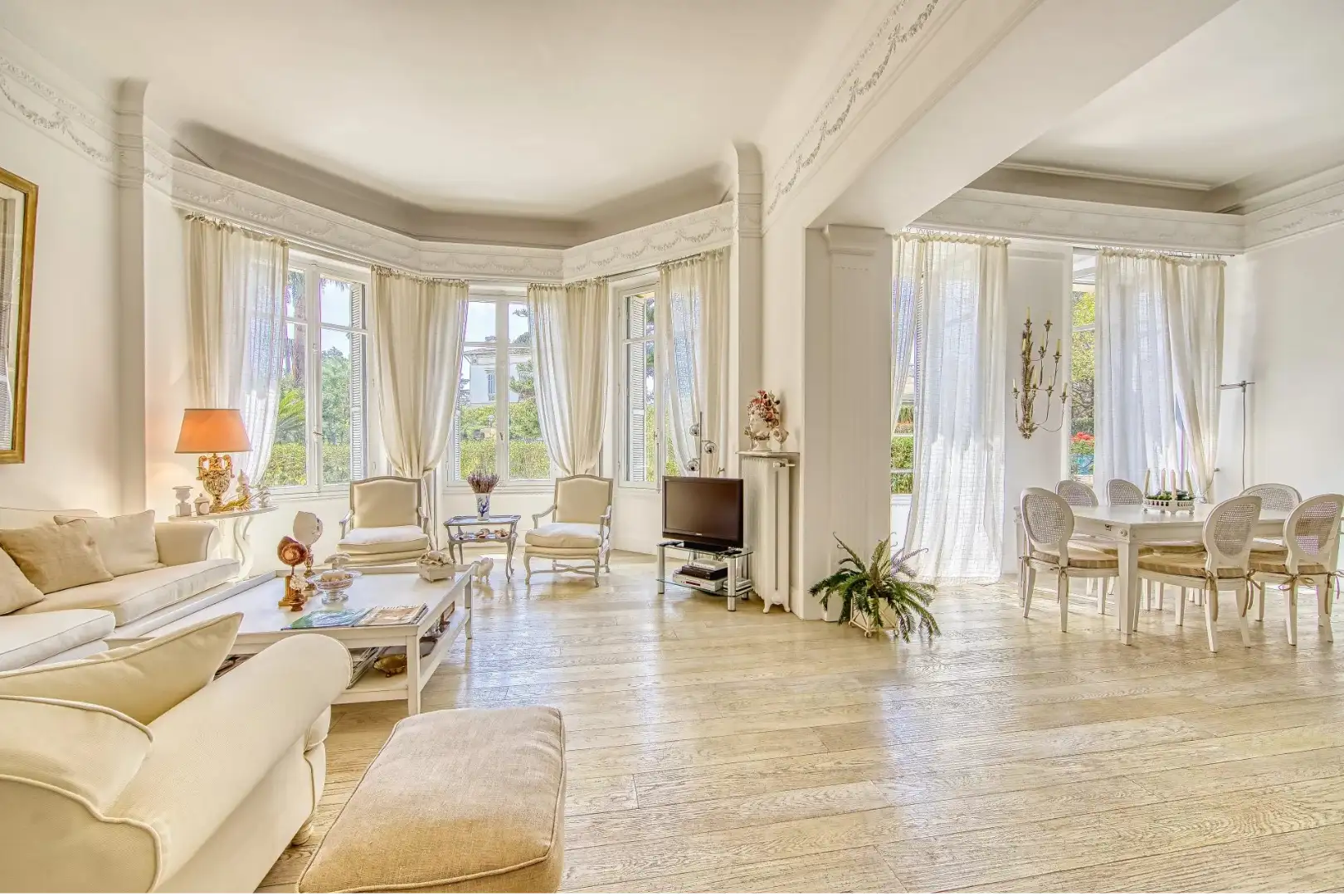 Grand séjour élégant avec salon et salle à manger attenante, meubles clairs, parquet et grandes baies vitrées avec rideaux blancs.
