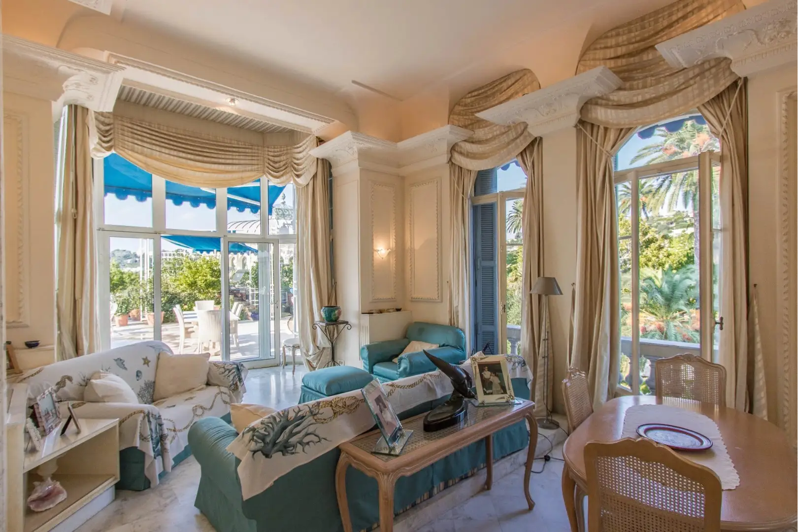 Salon raffiné avec canapés bleus, baies vitrées donnant sur une terrasse ensoleillée et rideaux dorés drapés.