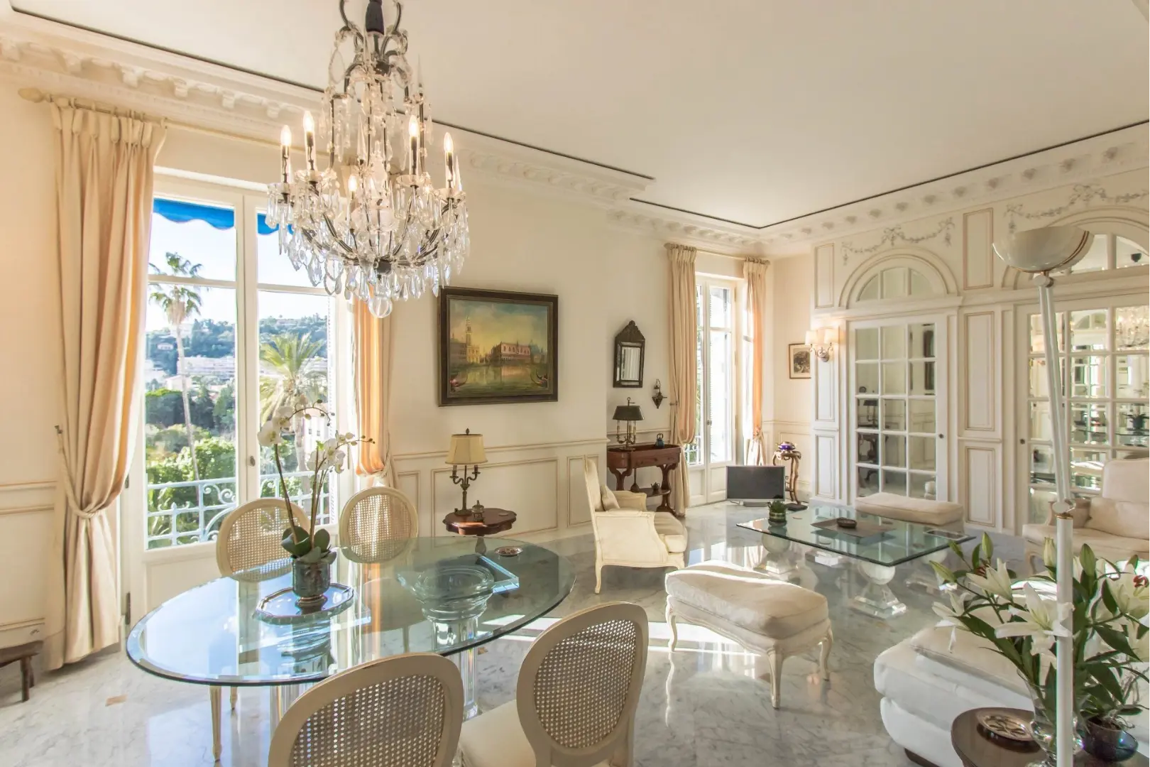 Séjour spacieux avec table en verre, chandelier central, moulures murales et vue sur jardin depuis de grandes fenêtres.