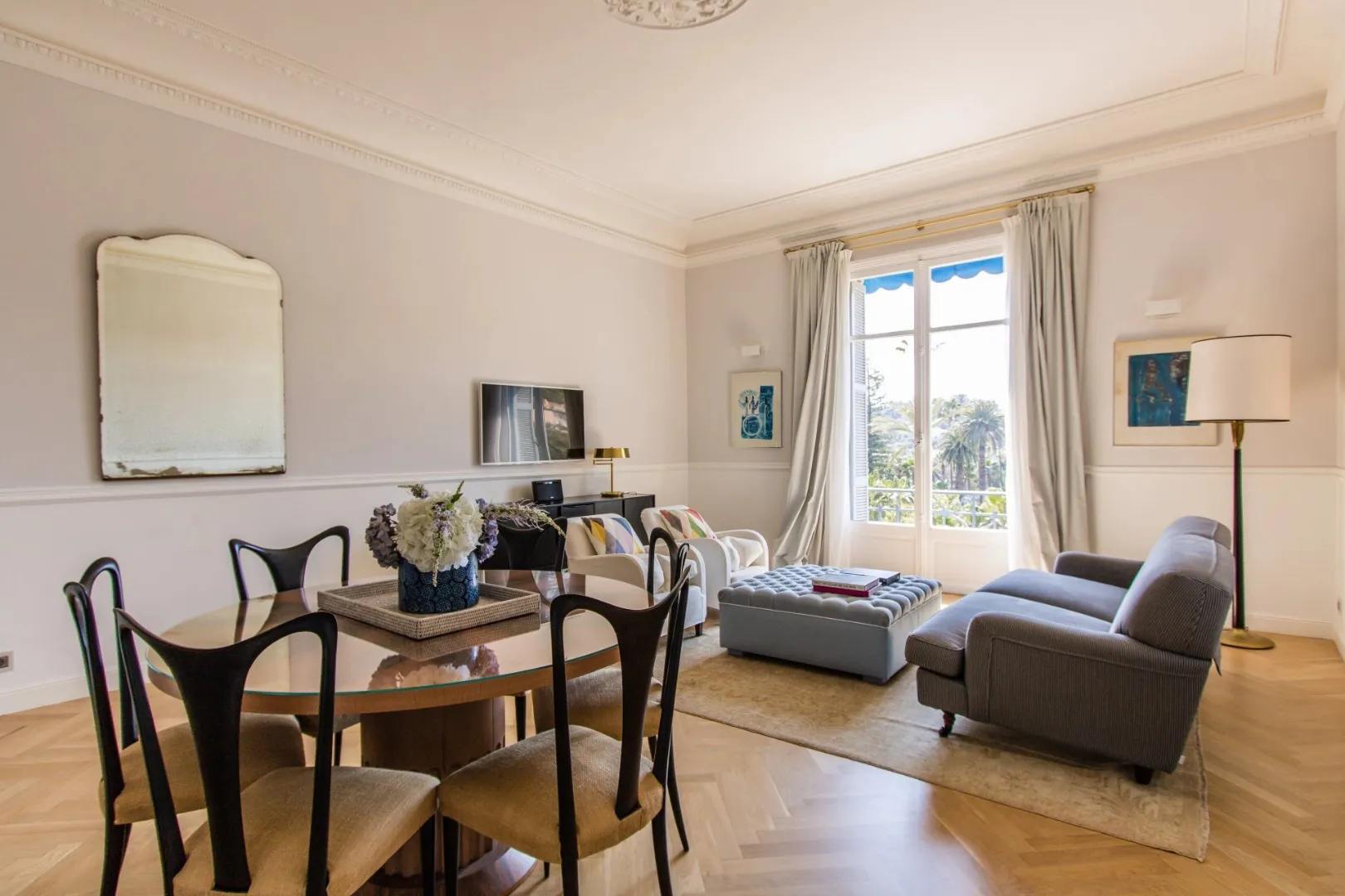 Salon moderne avec canapé gris, table basse rembourrée, coin salle à manger et grande fenêtre donnant sur un paysage tropical.