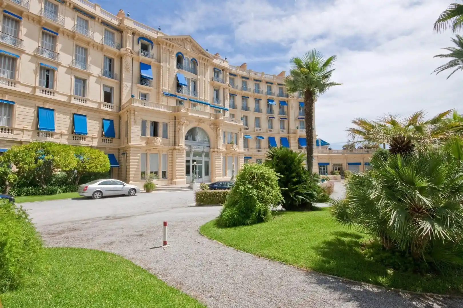 Façade incurvée d’un bâtiment Belle Époque couleur crème, volets et auvents bleus, allée gravillonnée bordée de pelouses et de palmiers, voitures en stationnement.